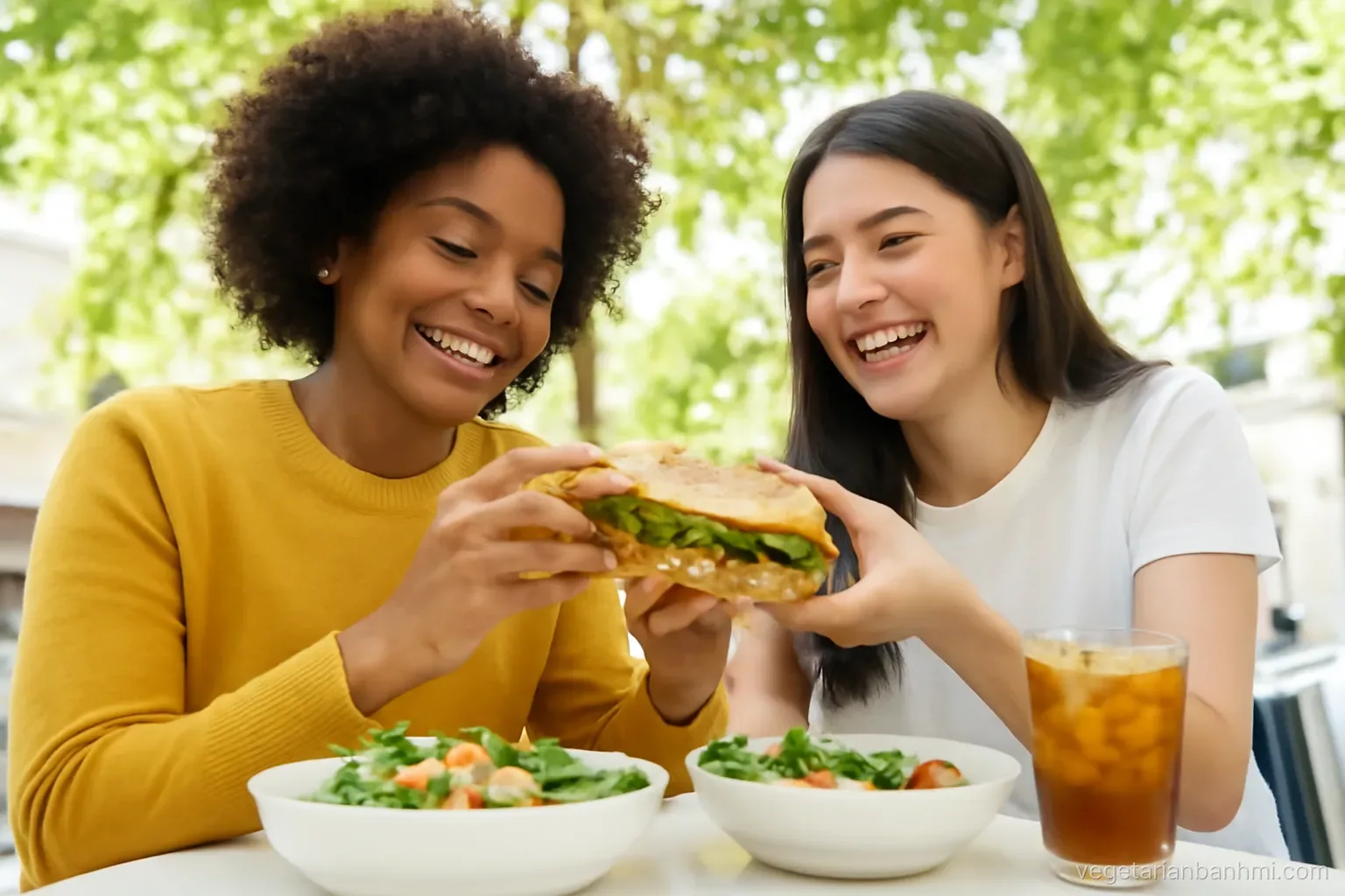 Two friends of different ethnicities share a vegan Vietnamese baguette sandwich split lengthwise with salad bowls and herbal iced tea at an outdoor cafe under trees, low angle.