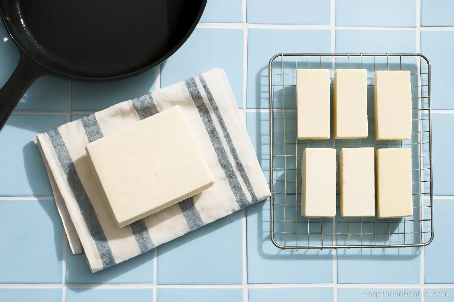 Bird's eye view of a tofu block in a striped linen towel under a cast iron pan on a light blue tiled counter, tofu slabs on a rack, crisp daylight