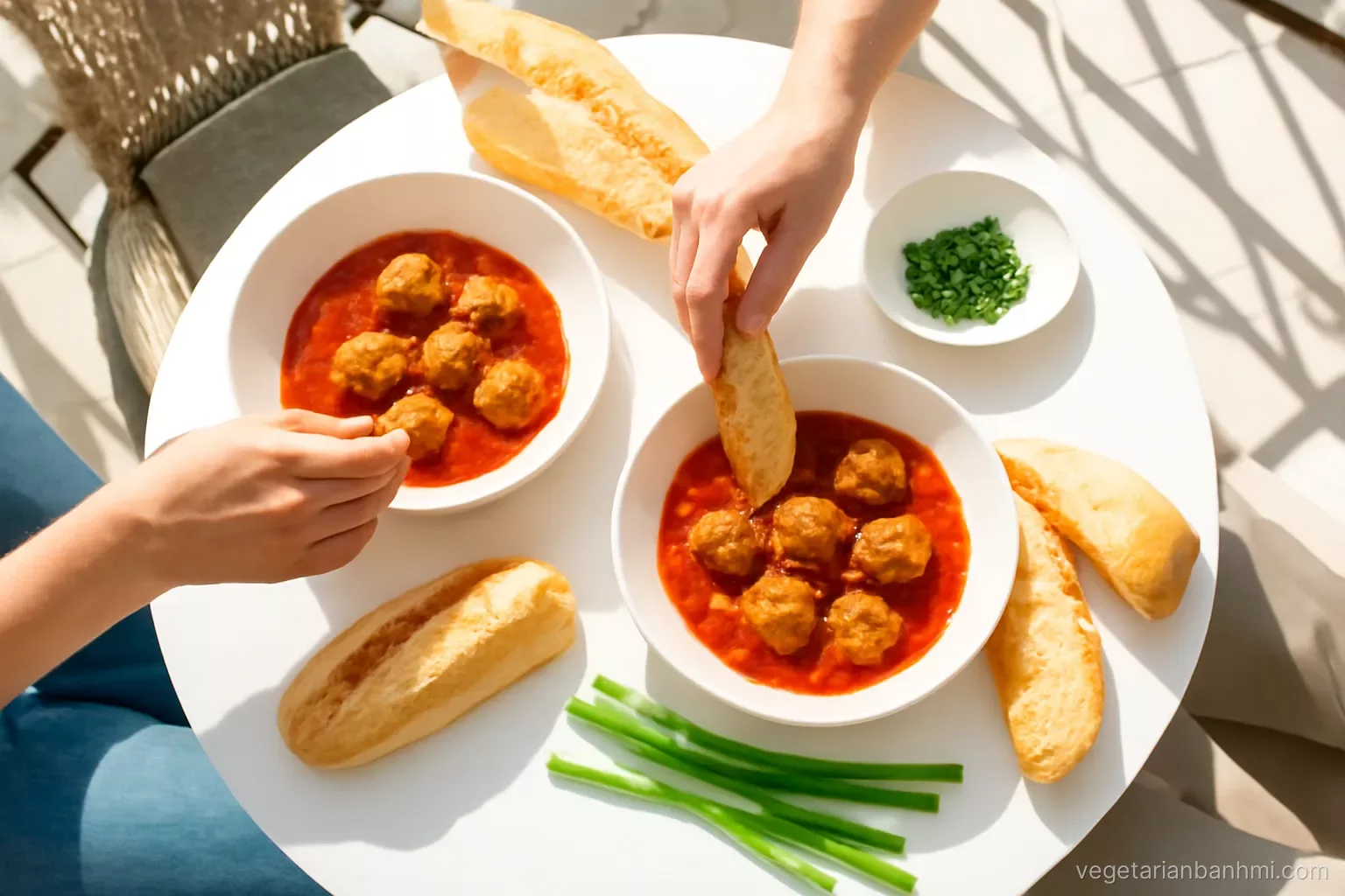 Overhead view of a sunny balcony breakfast table with bowls of tofu meatballs in red tomato sauce, crusty Vietnamese baguettes split lengthwise, scallions, two people reaching in.